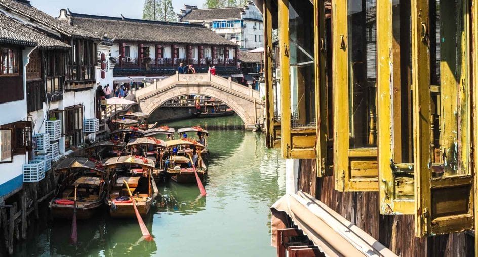 A stone arch bridge over a canal in Zhujiajiao, with flat-bottomed tourist boats moored along the water below whitewashed houses.