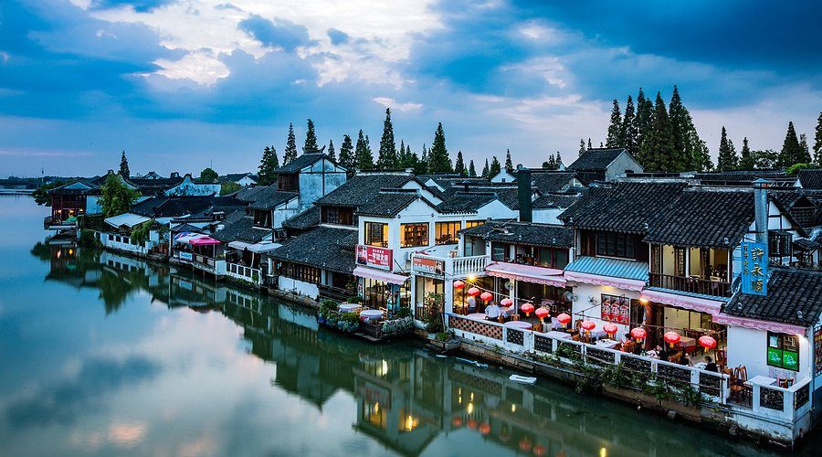 A row of whitewashed, black-tiled waterfront houses along a broad canal in Zhujiajiao under a blue sky, awnings and red lanterns at the water.