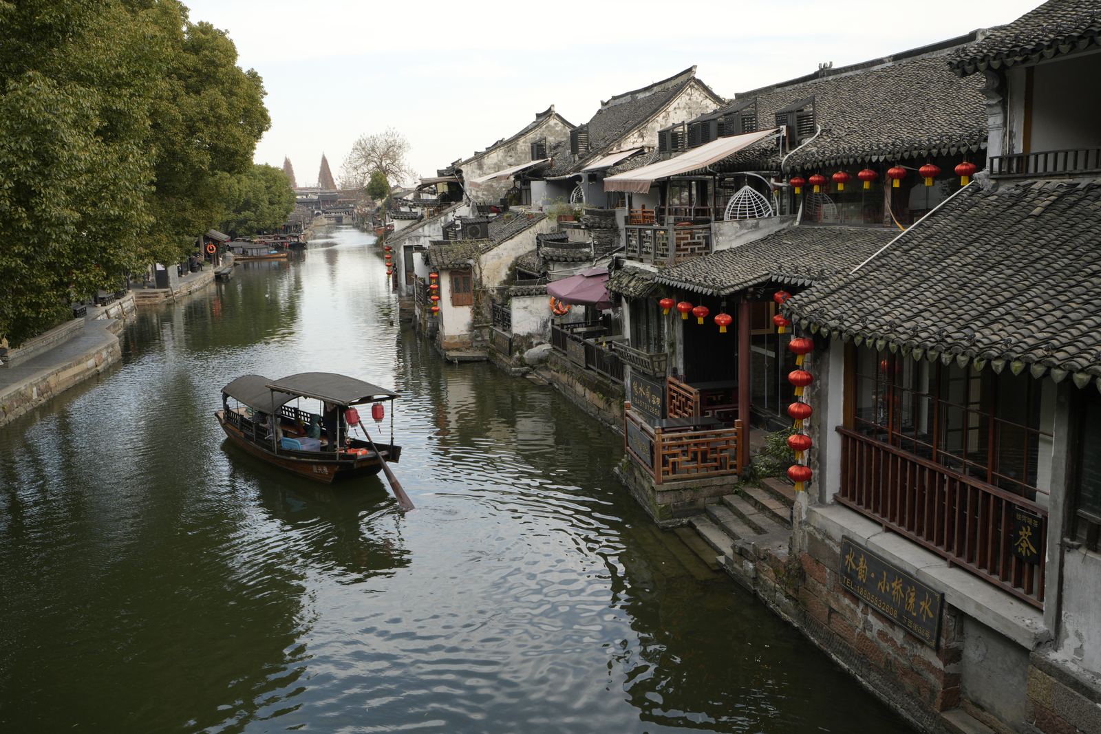 A canal in Xitang in daylight, a small covered boat on the water between timber houses hung with red lanterns.