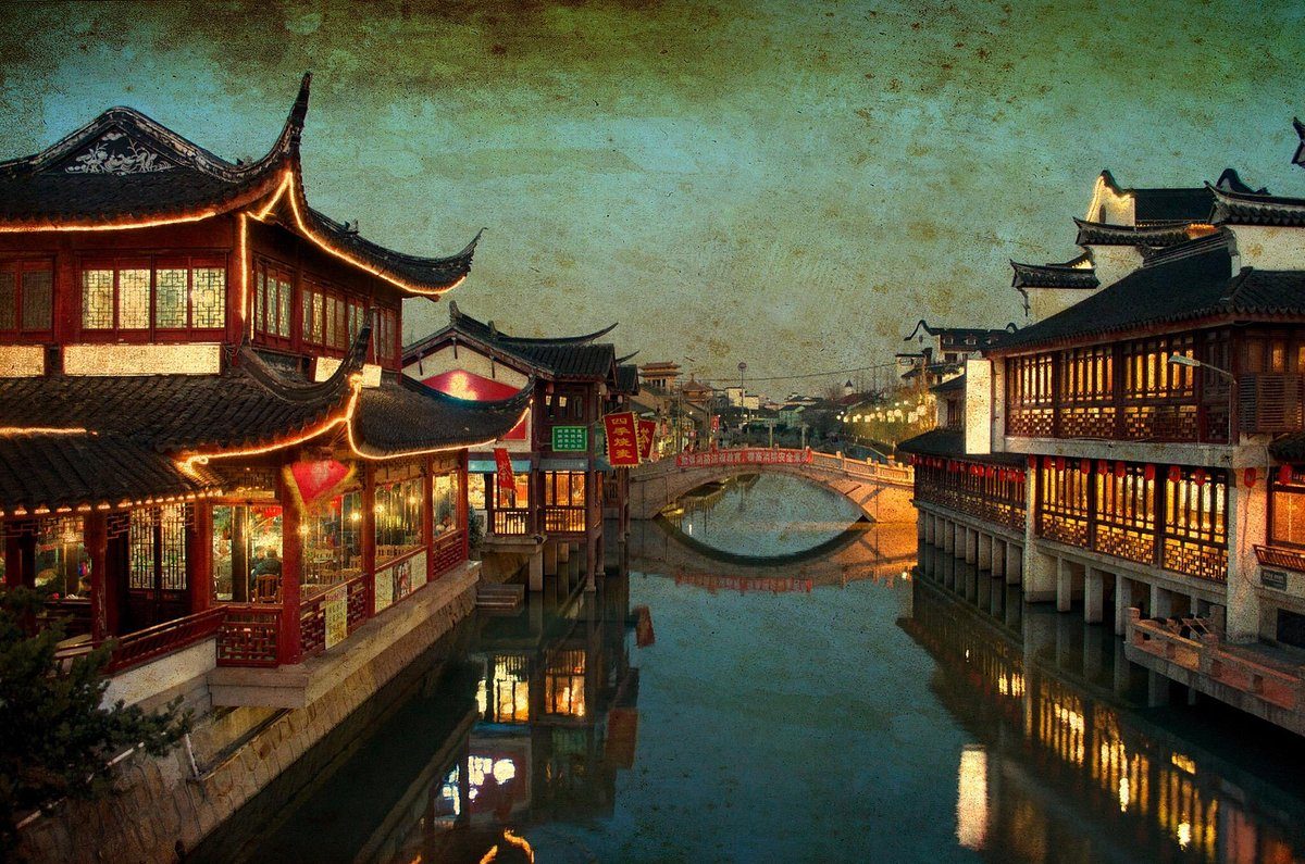 Qibao Old Street at night, lit shopfronts and upturned eaves reflected in the canal, with a low stone bridge in the distance.