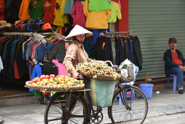 A street vendor pushing a bicycle loaded with baskets of fruit past clothing shopfronts in Hanoi's Old Quarter.