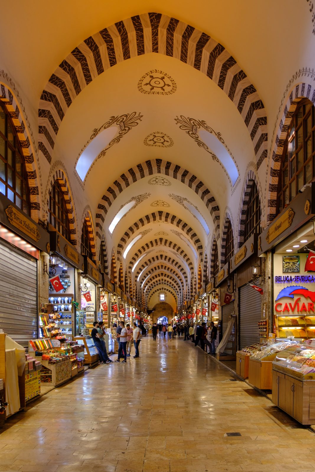 The vaulted arcade of the Spice Bazaar, shopfronts and Turkish flags running the length of the hall under painted arches.