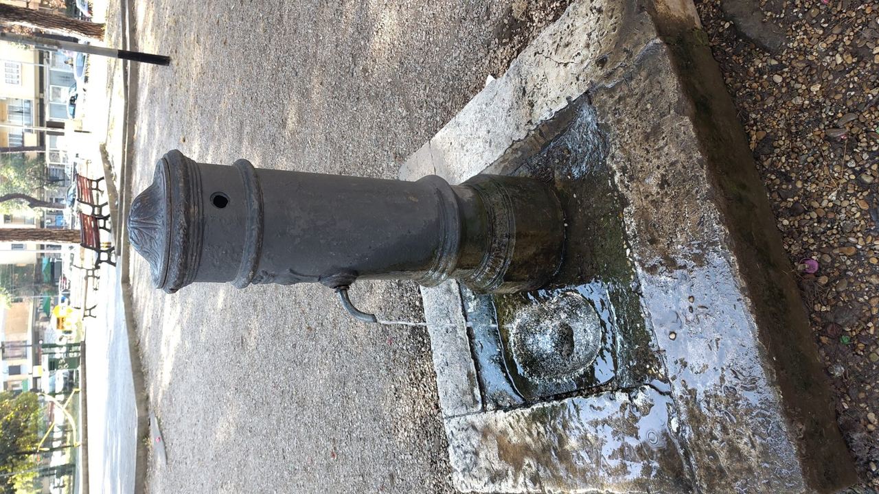 Water running continuously from the curved spout of a cast-iron nasone drinking fountain onto the wet stone of a Rome street.