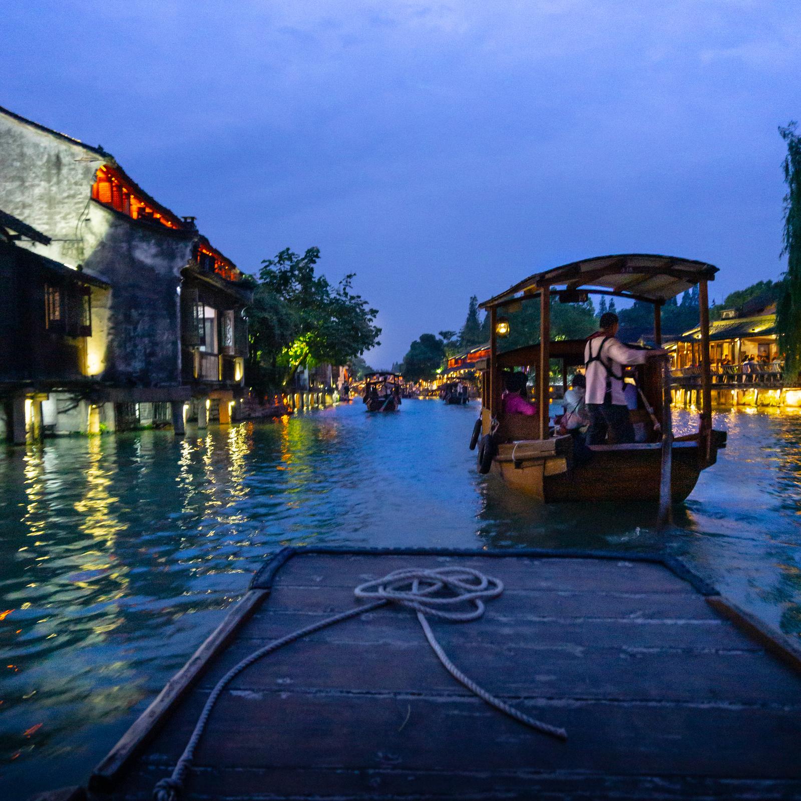 A wooden boat on a Wuzhen canal at blue hour, lamplight from the timber buildings reflecting in the water.