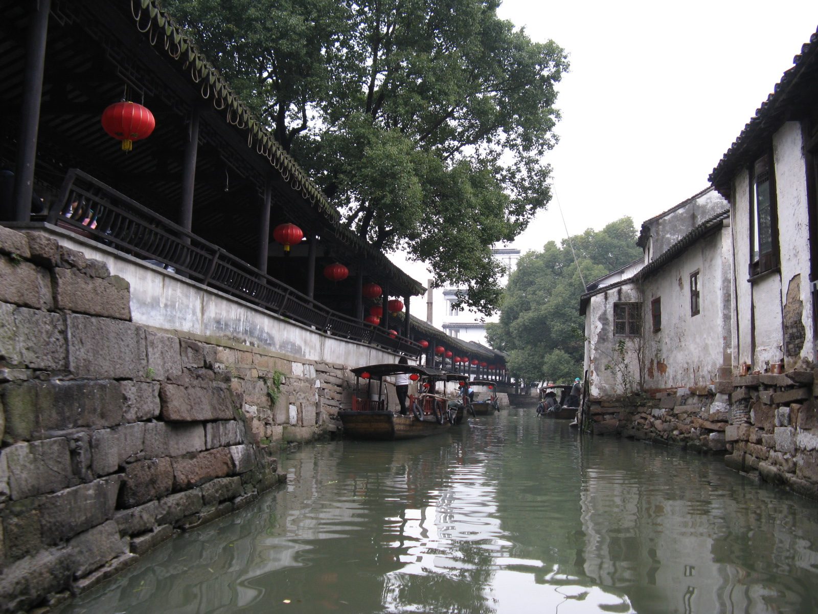 A stone-embanked canal in Jinxi running under a lantern-hung covered walkway, whitewashed houses on the far bank.
