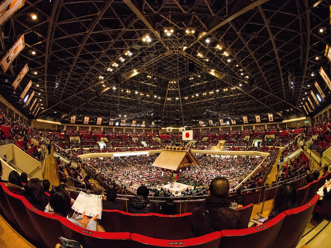 The packed interior of the Ryōgoku Kokugikan during a grand sumo tournament, with the roof suspended over the ring