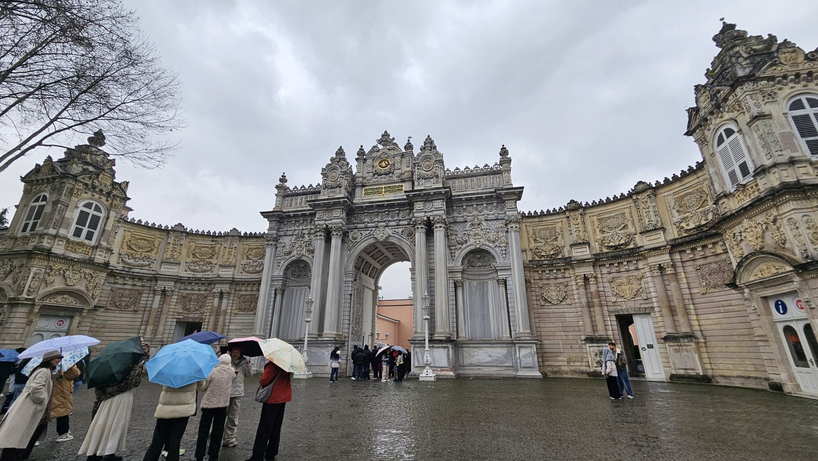The ornate carved gateway of Dolmabahçe Palace with its clock above the arch, visitors sheltering under umbrellas on the wet forecourt.
