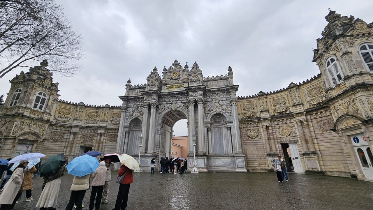 The ornate carved gateway of Dolmabahçe Palace with its clock above the arch, visitors sheltering under umbrellas on the wet forecourt.