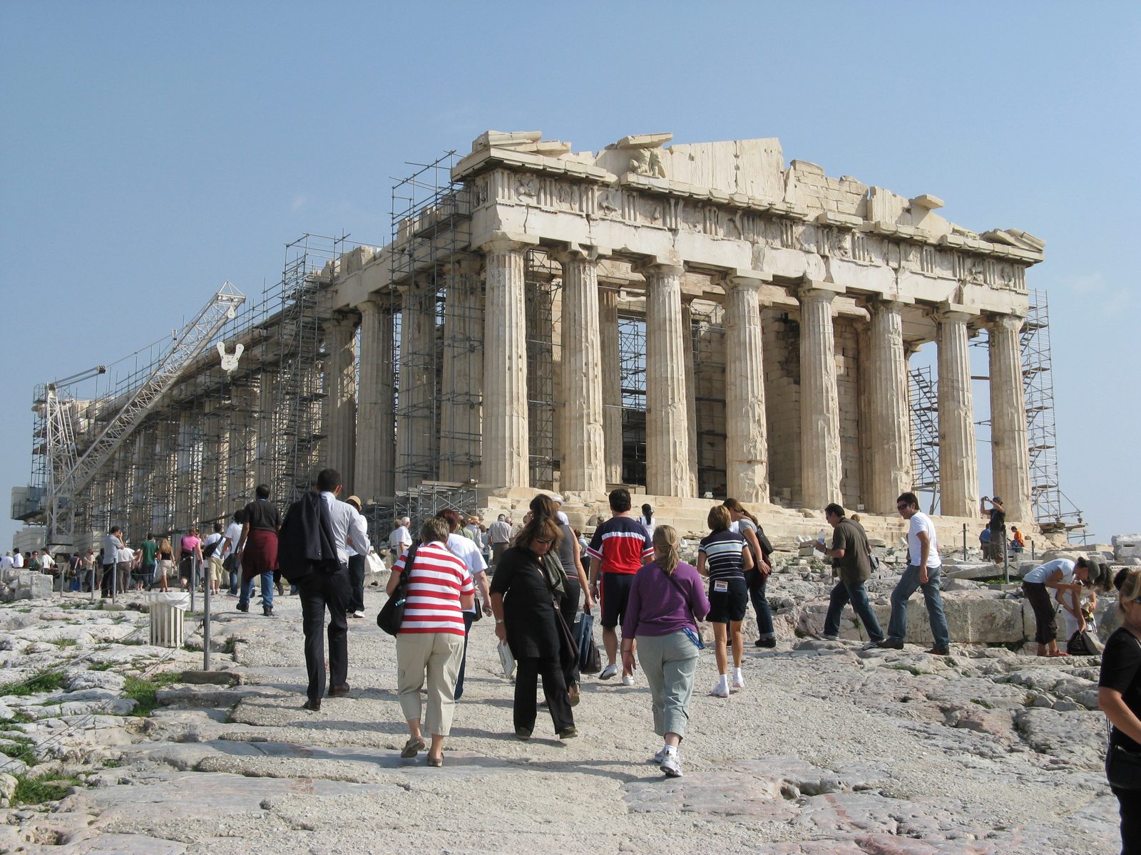 Visitors walking across the bare, sunlit rock of the Acropolis in front of the Parthenon, with no shade anywhere on the hilltop.