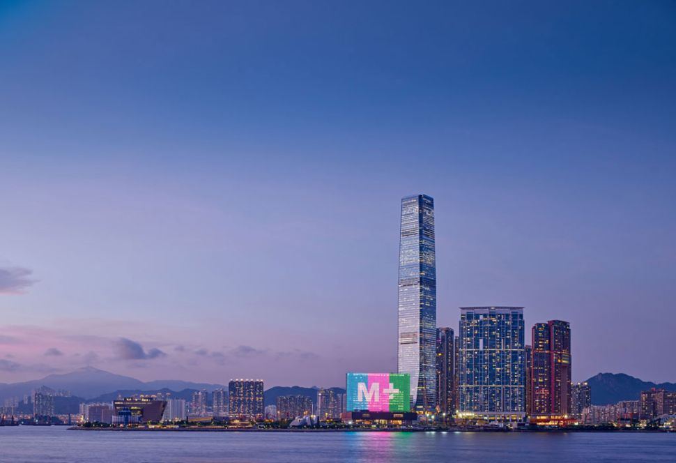 The M+ museum tower on the West Kowloon waterfront at dusk, its logo lit on the building